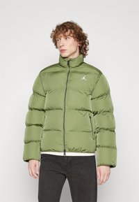 Giacca imbottita color verde oliva con colletto alto, chiusura con zip e motivo a quadri. Presenta un logo bianco sul petto sinistro e tasche laterali.