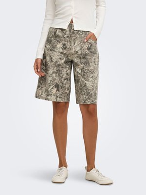 Personne portant un short à motifs gris et beige jusqu'aux genoux, un haut blanc à manches longues boutonné et des baskets blanches décontractées, debout avec une main dans la poche.