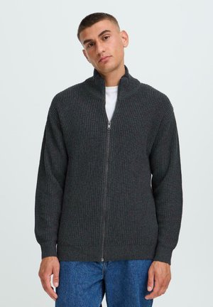 Dunkelgrau gerippter Strick-Zip-Hoodie mit hohem Kragen, langen Ärmeln und einer strukturierten Oberfläche. Über einem weißen Rundhalsausschnitt-T-Shirt getragen.
