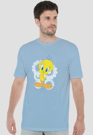 Mann trägt hellblaues T-Shirt mit einem großen bunten Cartoon-gelben Vogel und weißem floralem Hintergrund vorne, schaut leicht nach rechts.
