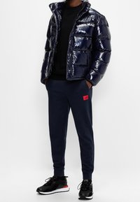Marineblå puffjakke med blank finish, sort sweatshirt, marineblå sweatpants med rød logo-patch, og sorte træningssko.