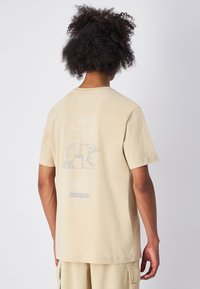 Camiseta de algodón beige con un gráfico de oso y texto en la parte posterior. Manga corta y corte relajado con un escote redondo.