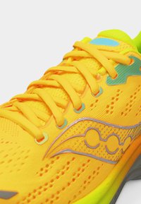 Φωτεινό κίτρινο παπούτσι τρεξίματος με πορτοκαλί κορδόνια, μπλε και ασημί λογότυπα, και διαπνέον ύφασμα από δίχτυ στην επάνω επιφάνεια.