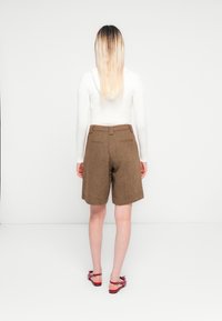 Frau mit langen Ombre-Haaren, die einen weißen cropped Hoodie, braune knielange Shorts und rote flache Sandalen trägt und vor einer weißen Wand steht.