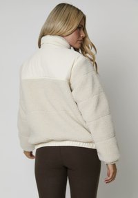 Mujer con cabello rubio largo, vestida con una chaqueta de sherpa color crema con la parte superior trasera acolchada y pantalones ajustados marrón oscuro, de espaldas.