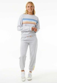 Joven mujer con sudadera gris claro y joggers a juego con rayas azules, naranjas y amarillas, de pie y sonriendo.