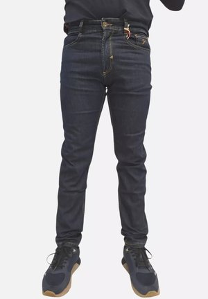 Persona che indossa jeans aderenti blu scuro e scarpe da ginnastica nere con suola beige, in piedi davanti a uno sfondo chiaro e uniforme.