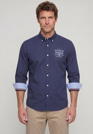 Hombre que lleva una camisa azul marino abotonada con las mangas remangadas y pantalones beige, de pie frente a un fondo claro y liso.