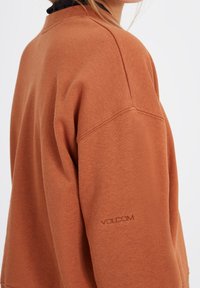Sweat-shirt marron à coupe décontractée, col rond côtelé et logo "VOLCOM" brodé sur la manche gauche. Tissu doux avec une texture lisse.