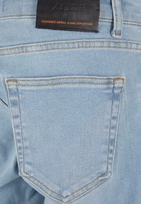 Poche arrière d'un jean en denim bleu clair avec surpiqûres orange et une étiquette de marque en cuir noir avec texte orange embossé.