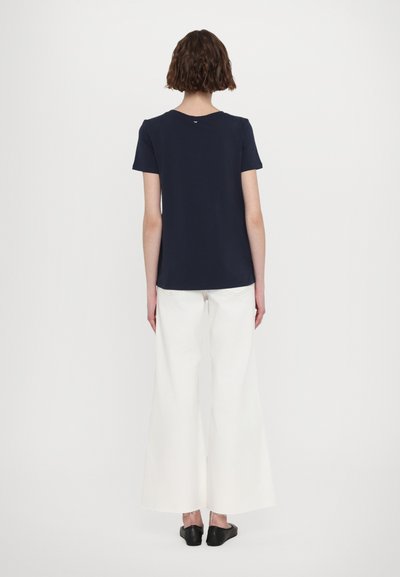 T-shirt bleu marine à manches courtes avec un col rond, associé à un pantalon large blanc et des chaussures noires à enfiler, vu de dos.