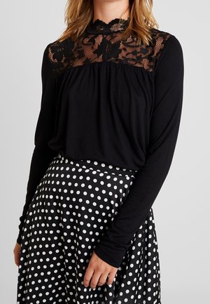 Vrouw draagt een zwarte lange mouwen top met doorzichtige fleurige kanten inzet en een zwarte hoge taille rok met witte stippen.