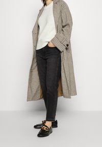 Abrigo largo de cuadros en tonos neutros, suéter de punto blanco, jeans oscuros y mocasines negros con un detalle de cadena dorada.