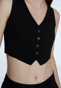 Gilet nero corto con scollatura a V, cinque bottoni neri e una silhouette aderente. Realizzato in un tessuto morbido con un design sartoriale.