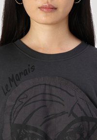 T-shirt grigio scuro con un design prominente di un volto stampato e dettagli di cucitura, testo "LeMarais" in un font audace sul davanti.