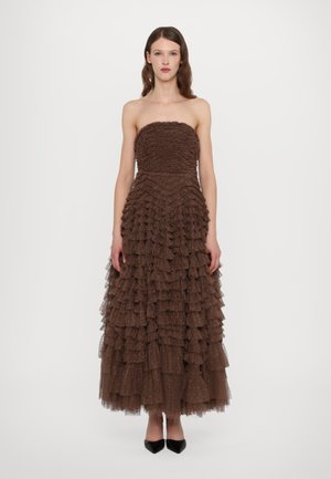 Robe marron sans bretelles faite de couches de volants, avec un buste texturé et une jupe fluide. Accompagnée de chaussures noires à bout pointu.