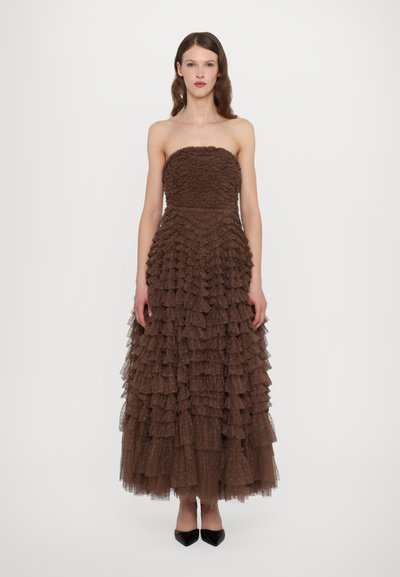 Robe marron sans bretelles faite de couches de volants, avec un buste texturé et une jupe fluide. Accompagnée de chaussures noires à bout pointu.