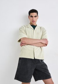 Light green, ribbed short-sleeve shirt with a pocket, paired with dark gray corduroy shorts. Model stands with arms crossed against a plain background.