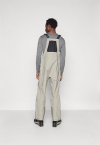 Beige skidsoverall med en svart bakficka, justerbara axelremmar, dragkedjor och förstärkta nedre ben, utformade för utomhusaktiviteter.