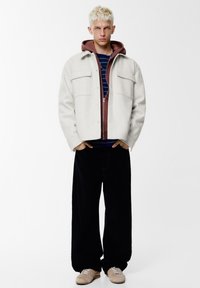 Off-white wollen jas met twee borstzakken over een bordeauxrode hoodie met rits, gedragen over een marineblauw gestreept shirt, gecombineerd met zwarte broek met wijde pijpen en beige sneakers.