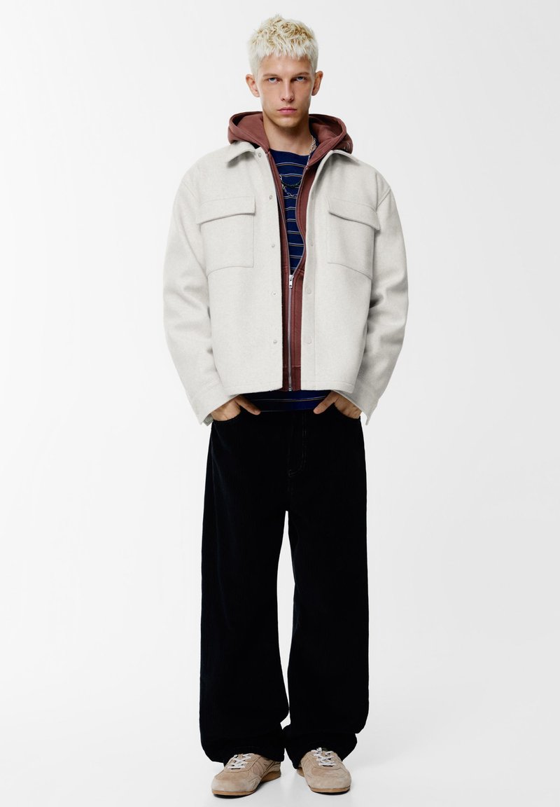 Off-white wollen jas met twee borstzakken over een bordeauxrode hoodie met rits, gedragen over een marineblauw gestreept shirt, gecombineerd met zwarte broek met wijde pijpen en beige sneakers.