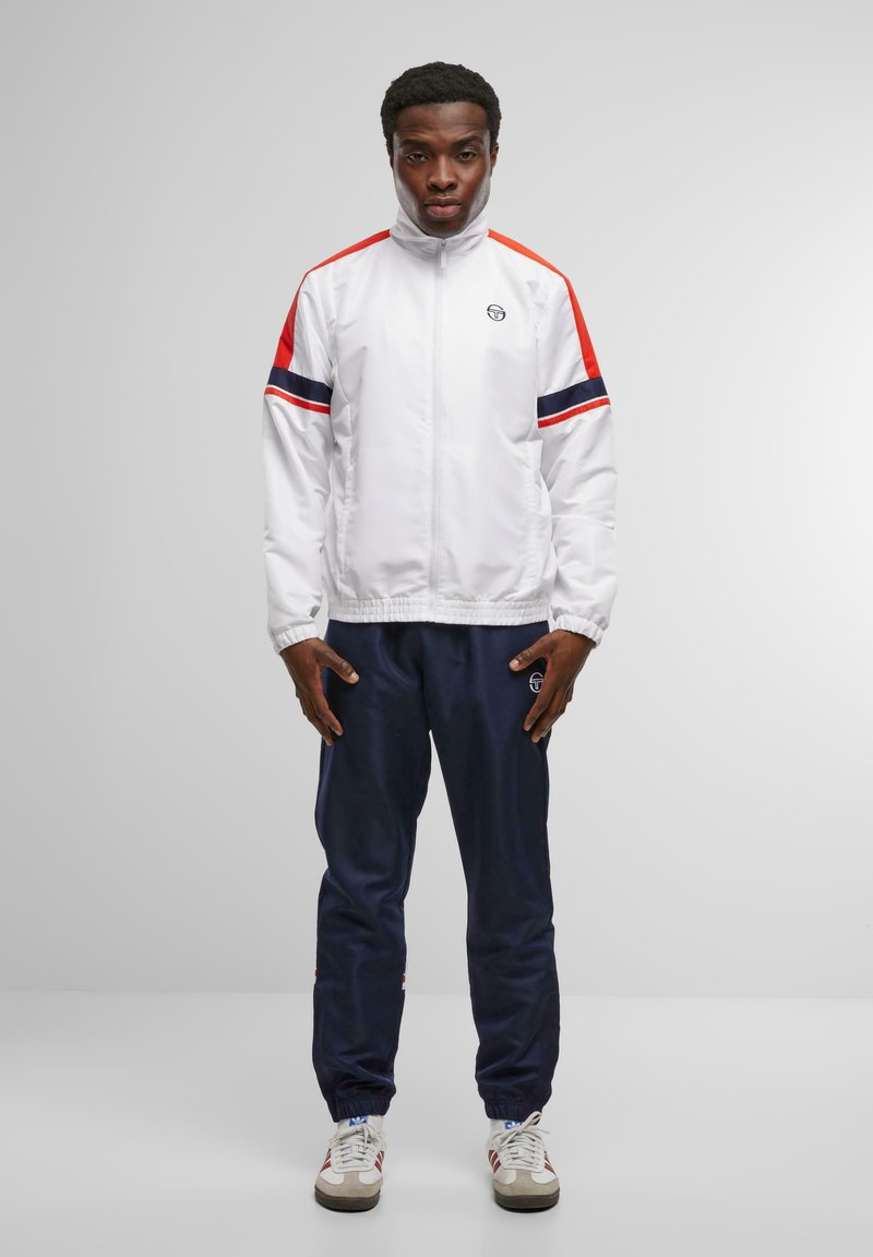 White athletic jacket with red and navy stripes, zip closure, paired with navy pants and white sneakers. Features a logo on the chest.