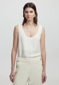 White sleeveless top with a deep scoop neckline, made of a smooth fabric, paired with cream trousers featuring gold button accents on the side.
