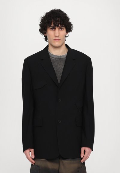 Schwarzer, maßgeschneiderter Blazer mit zwei vorderen Taschen, zwei Knöpfen, strukturierten Revers und lockerer Passform, getragen über einem grauen T-Shirt.