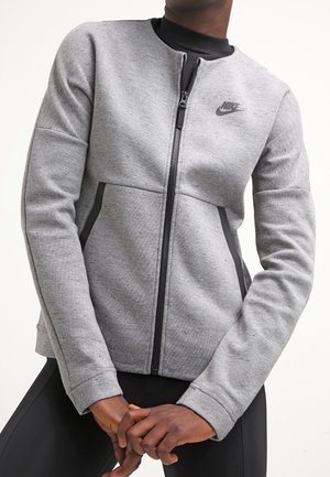 Person iført en lysegrå Nike jakke med lynlås med sorte detaljer og sorte træningsbukser, stående med hænderne foldet foran.