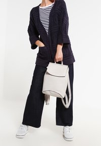 Ljusgrå, strukturerad ryggsäck med lockstängning, justerbara remmar och slät yta. Bärs med en marinblå kofta och randig topp.