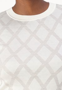 Acercamiento de una persona que lleva un suéter de punto blanco con un patrón de diamantes en beige claro y un cuello redondo con canalé.