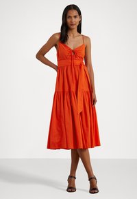 Vestido maxi em algodão laranja brilhante, com corpete ajustado, detalhe de laço e design de saia em camadas. Modelo usa sandálias pretas de tiras.