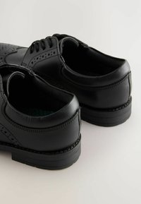 Des chaussures en cuir noir présentent une texture lisse, des détails en brogue et une forme arrondie. Elles possèdent une semelle robuste et des accents de couture contrastants.
