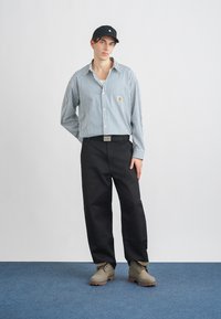 Camisa abotonada de rayas azul y blanca, pantalones negros y botas grises. Gorra negra, con logo prominente en la camisa. Diseño sencillo, ajuste informal.