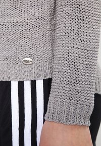 Szary dzianinowy sweter z teksturowanym wzorem, ściągaczami na rękawach i metalową metką z logo. Do tego czarne spodnie z białymi paskami.