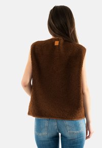 Gilet in pile marrone con superficie testurizzata, design senza maniche e un'etichetta vicino al collo. Abbinato a jeans blu con tasche posteriori.