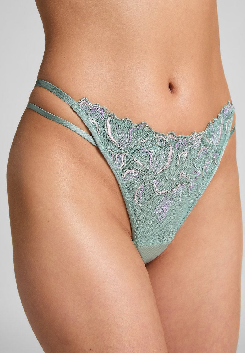 Grüner Spitzenstring mit floralen Stickereien in Pink und Lavendel, mit dünnen Trägern und transparenter Textur, die filigrane Designdetails hervorhebt.