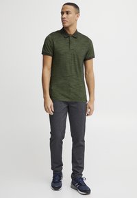 Mørkegrøn polo-shirt med en struktureret overflade, der har en to-knaps placket og kontrastfarvet krave, parret med mørkegrå slim-fit bukser og sneakers.