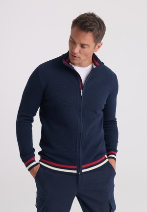 Homme en pull zippé texturé bleu marine avec poignets et ourlet rayés rouge, blanc et bleu, portant un pantalon bleu marine, regardant vers le bas à sa gauche.