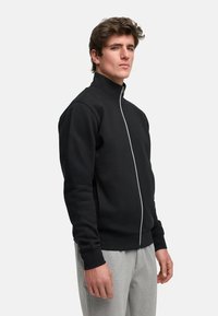 Svart zip-up hettegenser med høyt krage, med en kontrasterende glidelås. Stoffet ser glatt ut, og det er kombinert med lysegrå bukser.