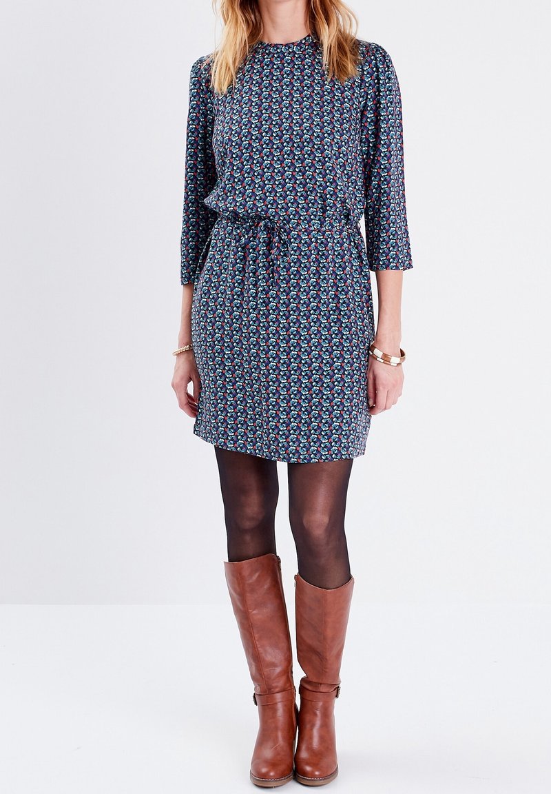 Kurzes Kleid aus dunklem Stoff mit buntem Blumenmuster, tailliert in der Taille. Ein Paar braune knielange Stiefel vervollständigt den Look.