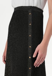 Una vista en primer plano de una mano reposando sobre una falda negra, elegante y brillante, con botones dorados que bajan por un lado.