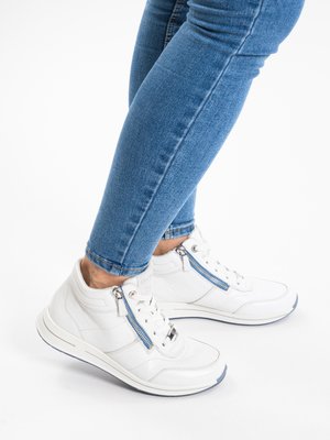 Person wearing blue skinny jeans and white high-top sneakers with side zippers, standing on a white background.