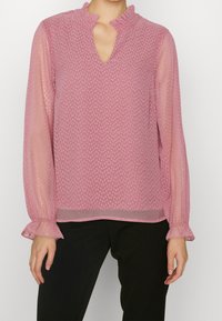 Blusa rosa trasparente con un motivo testurizzato, scollatura a volant e maniche lunghe con polsini arricciati. Taglio su misura con una leggera trasparenza.