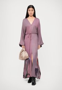 Abito lungo viola con scollatura a V profonda e maniche larghe. Presenta un motivo floreale e uno spacco frontale. Abbinato a una borsa morbida color beige.