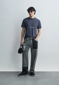 Γκρι γραφικό t-shirt με επιγραφή «KIDSUPER», σε συνδυασμό με γκρι τζιν patchwork με μαύρες και υφασμάτινες υφές. Μαύρα αθλητικά παπούτσια και μικρή τσάντα.