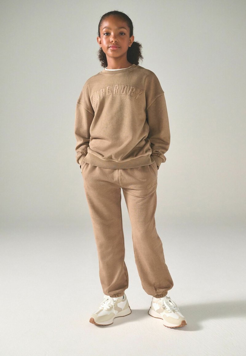 Brun bomullströja med präglad "SUPERDRY"-logotyp, matchad med joggers i samma färg och vita sneakers med beige detaljer.