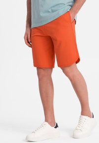 Shorts orange en tissu doux avec une texture lisse, dotés de poches latérales et d'un ourlet arrondi, accompagnés de baskets blanches.