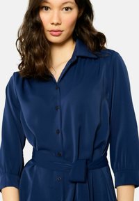 Robe chemise bleu marine en tissu satiné, dotée d'un col, d'une fermeture à boutons à l'avant et d'une ceinture à nouer à la taille. Manches trois-quarts avec poignets froncés.