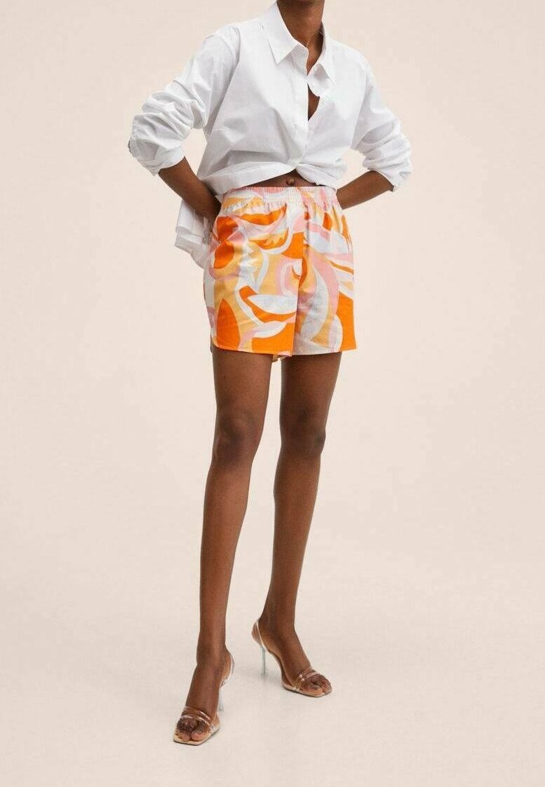 Personne portant une chemise blanche boutonnée avec les manches retroussées, un short à motifs multicolores orange et rose, et des chaussures à talons hauts transparentes.